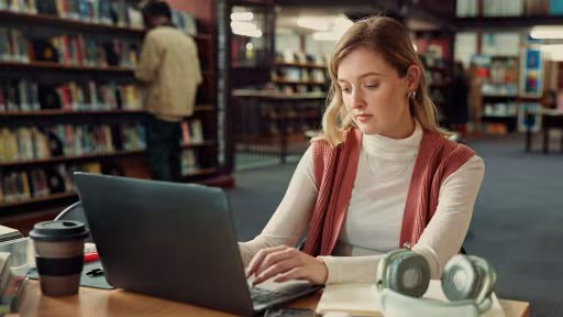 Junge Frau sitzt in einer Bibliothek und schreibt am Laptop