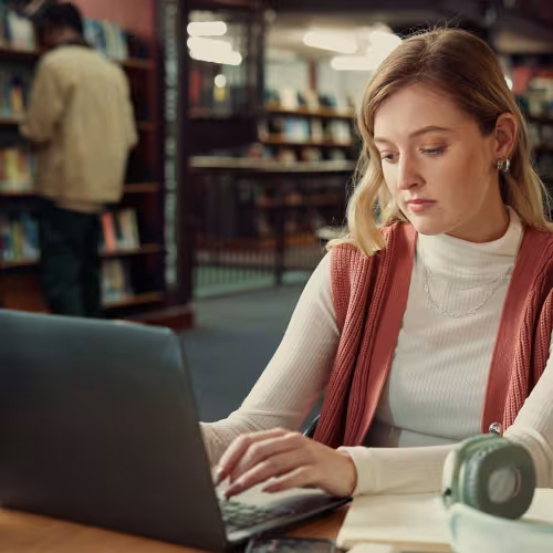 Junge Frau sitzt in einer Bibliothek und schreibt am Laptop